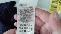 Metka ubraniowa trzymana w ręce zawiera skład tkaniny w wielu językach i zawiera symbole instrukcji pielęgnacji.