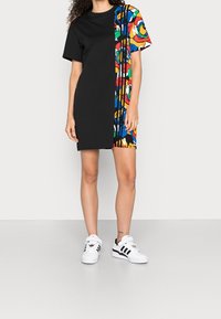 Černé krátké šaty s barevným květinovým vzorem na jedné straně. Vyrobené z bavlny, mají volný střih a kulatý výstřih. Bílé tenisky.