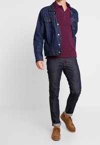 Džínová bunda s kapsami, vínová polo košile, tmavé džíny a hnědé kotníkové boty. Otočené manžety na džínách a viditelné švy.