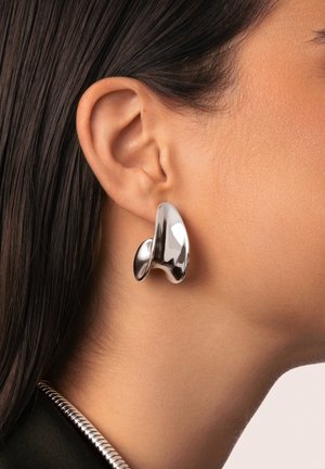 Boucle d'oreille sculpturale en argent métallique avec une forme fluide, présentant des surfaces lisses et réfléchissantes ainsi qu'un design abstrait.