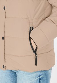 Beige quilted pufferjakke med en sort lynlås lomme og sorte trykknapper. Sammen med lyseblå denim.