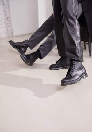 Zwarte leren combatlaarzen met ronde neuzen, vetersluiting en dikke rubberen zolen. De schoenen hebben witte stiksels en een gladde textuur.