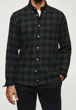 Homme portant une chemise à carreaux vert foncé et noir boutonnée sur un tee-shirt blanc, avec un pantalon noir, une main dans la poche.