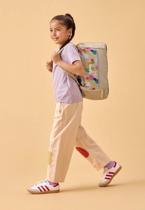Beige Rucksack mit buntem Punktedesign, helllila Hemd, beige Hose mit roten und gelben Flecken und weiße Turnschuhe mit roten Streifen.