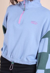 Femme portant un pull en polaire cropped bleu clair avec une fermeture éclair rose, un motif géométrique vert et bleu sur les manches, et un pantalon noir.