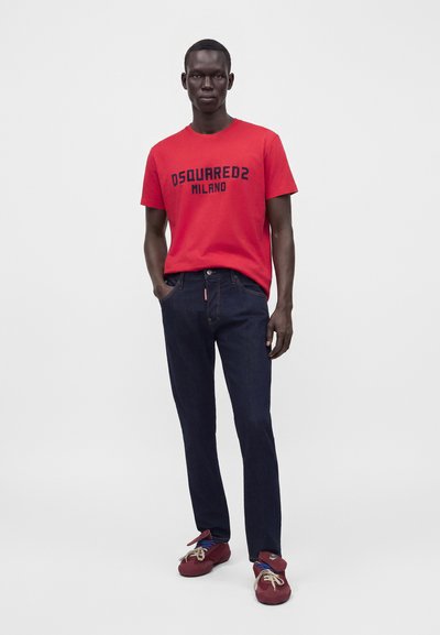 Άνδρας που φοράει κόκκινο T-shirt Dsquared2 Milano, σκούρο μπλε τζιν και κόκκινα sneakers, στέκεται με το ένα χέρι στην τσέπη μπροστά σε μονόχρωμο φόντο.