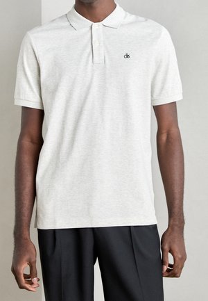 Mand iført lys grå kortærmet poloshirt med lille sort logo på brystet og sorte bukser, stående mod en ensfarvet baggrund.