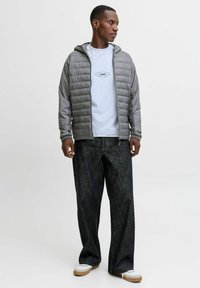 Grå, fyldt jakke med hætte over en lysegrå t-shirt med et "1990" grafisk tryk. Mørke, løstsiddende jeans og hvide sneakers fuldender looket.