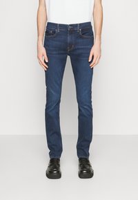 Jeans de mezclilla azul oscuro con ajuste slim, que cuentan con cinco bolsillos, pasadores para el cinturón y costuras en contraste. Llevados con zapatos negros.