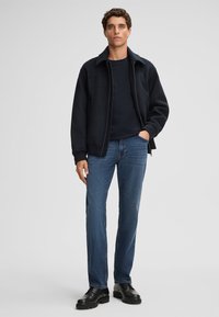 Veste zippée noire sur une chemise sombre, associée à un jean en denim bleu et des chaussures noires à lacets. Le modèle se tient droit contre un fond clair.