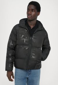 Schwarze Pufferjacke mit glänzender Textur und tonalen Logos, ausgestattet mit einem Reißverschluss vorn und einer Kapuze, getragen über einem schwarzen Shirt.