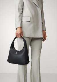 Czarna skórzana torebka z zaokrąglonym uchwytem i subtelnym złotym logo, w parze z jasnoszarym, dopasowanym garniturem, tworzącym elegancki wizualny kontrast.