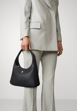 Czarna skórzana torebka z zaokrąglonym uchwytem i subtelnym złotym logo, w parze z jasnoszarym, dopasowanym garniturem, tworzącym elegancki wizualny kontrast.