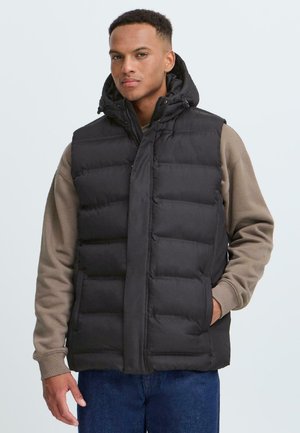 Black puffer vest with a hood, featuring a quilted design, zip front, and side pockets. Worn over a beige long-sleeve shirt.