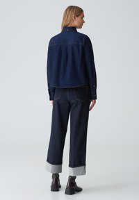 Giacca in denim scuro con un design cropped, caratterizzata da colletto e polsini con bottoni, abbinata a jeans in denim scuro a vita alta e gamba ampia con risvolti.