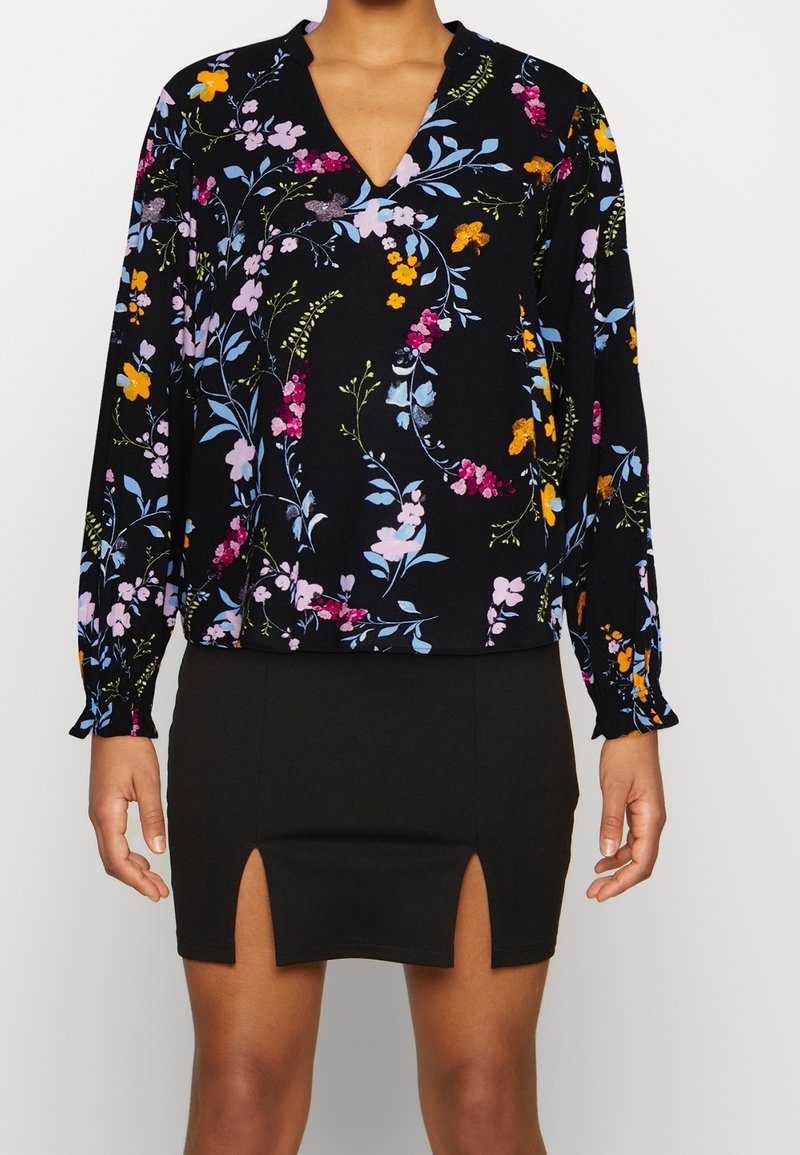 Blouse noire à imprimé floral coloré associée à une jupe noire avec deux fentes à l'avant.