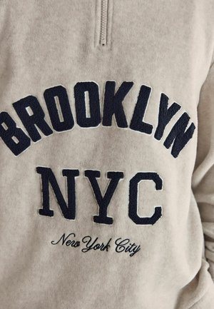 Beige Fleece-Pullover mit Viertelreißverschluss, auf dem in marinefarbener Stickerei der Text "BROOKLYN", "NYC" und "New York City" in verschiedenen Schriftarten und -stilen zu sehen ist.