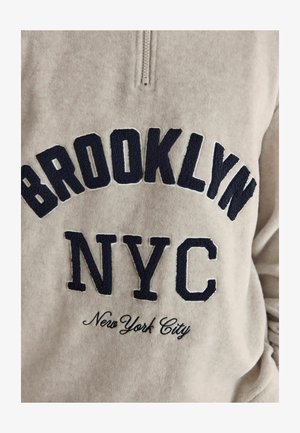 Bež fliissttage neljanda osa-üksik särk, millel on tumesinised broderitud tekstid: "BROOKLYN," "NYC," ja "New York City" erinevates fontides ja stiilides.