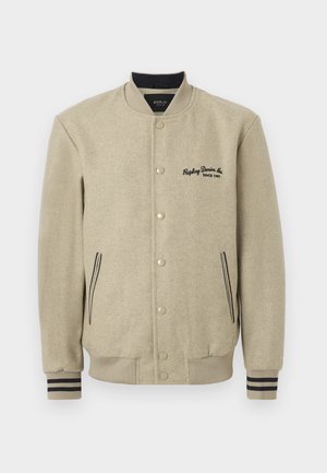 Beige színű egyetemi dzseki textúrált anyagból, pattintós gombokkal, hímzett logóval a mellrészen és kontrasztos csíkozott mandzsettával.