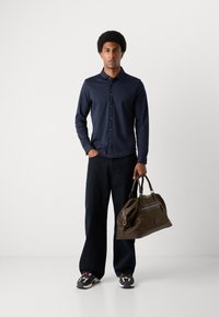 Marineblå langermet skjorte, mørk denim jeans og flerfargede joggesko. Bærer en romslig duffelbag i brun og oliven med en frontlomme med glidelås.