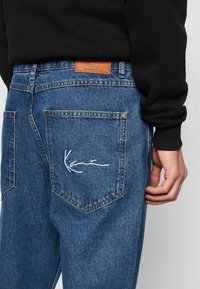 Denim jeans i mellemblå med en struktureret overflade. Har et lædermærke og hvidt broderet signatur på baglommen.