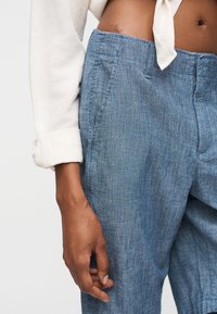 Κοντινό πλάνο ενός ατόμου που φοράει ανοιχτό μπλε λινό παντελόνι και λευκό μακρυμάνικο πουκάμισο δεμένο στη μέση, δείχνοντας το μέσο σώμα και το αριστερό χέρι.