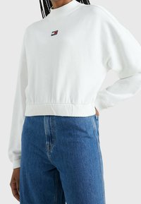 Sudadera blanca recortada con cuello alto, con un pequeño parche de logo en el pecho y puños acanalados. Combinada con vaqueros de mezclilla azules.