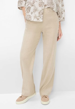 Person iført beige bukser med vide ben, en lys bluse med blomsterprint og lyse beige sneakers, stående med hænder delvist synlige.