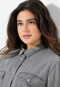 Camicia grigia con bottoni, dotata di due tasche sul petto e realizzata in tessuto testurizzato. Presenta un colletto e bottoni a pressione bianchi per la chiusura.