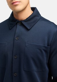 Mørkeblå skjorte med knapper, der har et tekstureret design, to forlommer, kravehals og sorte knapper. Glat stoffinish.