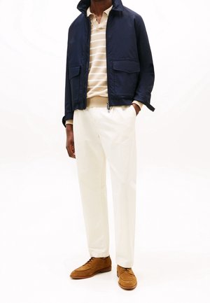 Homme portant une veste bleu marine, un pull rayé beige, un pantalon blanc et des chaussures de ville marron sur un fond blanc.