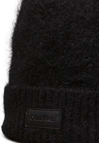 Berretto di maglia nera con una texture pelosa, caratterizzato da un risvolto a coste e un'etichetta in pelle con la scritta "Calvin Klein" in rilievo.