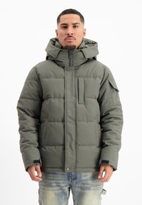 Olijfgroene pufferjack, met een hoge kraag, een rits aan de voorkant, grote zakken en een gestikte textuur. Gemaakt over lichtblauwe jeans.