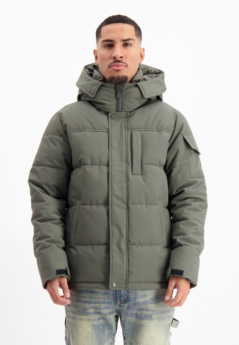 Olijfgroene pufferjack, met een hoge kraag, een rits aan de voorkant, grote zakken en een gestikte textuur. Gemaakt over lichtblauwe jeans.