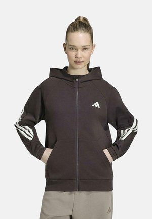 Donna che indossa una felpa nera con zip, strisce bianche sulle maniche e logo sul petto, con le mani nelle tasche, in piedi davanti a uno sfondo semplice.