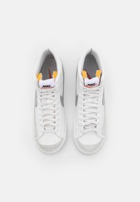 Zapatillas Nike de caña alta en cuero blanco con acentos de ante gris. Presenta una lengüeta acolchada, cordones blancos y un detalle amarillo en el cuello.