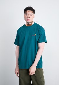 Teal cotton t-shirt with a crew neck, featuring a small embroidered logo in yellow and red on the left chest. Worn with olive cargo pants.