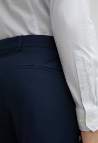 Weißes Hemd mit einem dezenten blauen Muster und geknöpften Manschetten, kombiniert mit dunkelblauen strukturierten Hosen mit einer seitlichen Tasche.