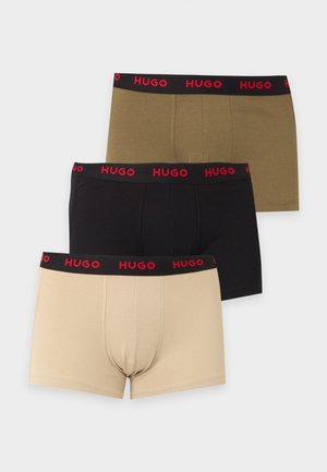 Atletický tmavšej pokožky muž stojaci bosý, oblečený v čiernych boxerkoch s červeným pásom s logom "Hugo" na bielom pozadí.