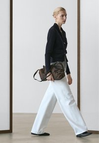 Donna che cammina al chiuso indossando una giacca blu navy, pantaloni bianchi a gamba larga, scarpe nere e portando una borsa a tracolla in pelle marrone.