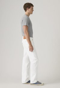 Νέος άντρας σε προφίλ, φορώντας γκρι μπλούζα, λευκά τζιν και μπλε αθλητικά παπούτσια, με απλό φωτεινό φόντο.