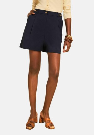 Vrouw draagt marineblauwe op maat gemaakte shorts, beige top, gestapelde houten armbanden en bruine sandalen met hak en gekruiste bandjes.