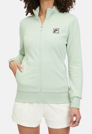 Osoba ubrana w jasnozieloną kurtkę zapinaną na suwak z logo na piersi i białe spodenki, z jedną ręką w kieszeni i kręconymi włosami.