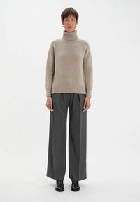 Un pull en laine beige avec col roulé et poignets côtelés, associé à un pantalon large à rayures grises et des chaussures noires, se tenant contre un fond blanc.