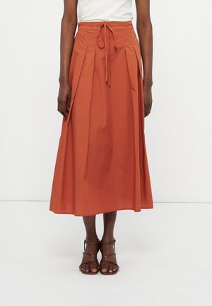 Mujer con una falda midi color óxido con pliegues y una cinturilla atada, combinada con sandalias marrones de tiras y una camiseta blanca.