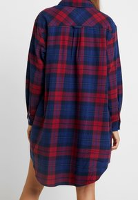 Röd och blå rutig skjortklänning med långa ärmar, knäppning vid ärmslut och rundad nederkant. Har en ryggås och avslappnad passform.