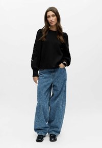 Svart genser med pufferm og blomsterformede utskjæringer, kombinert med vide, lyseblå mønstrede denimjeans og svarte sko.