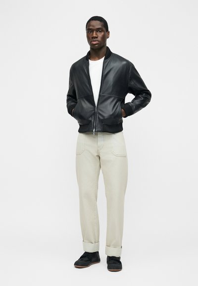 Άνδρας που φοράει μαύρο δερμάτινο μπουφάν τύπου bomber, λευκό μπλουζάκι, μπεζ παντελόνι με γυρισμένα μανίκια και μαύρα αθλητικά παπούτσια, στέκεται με τα χέρια στις τσέπες σε λευκό φόντο.