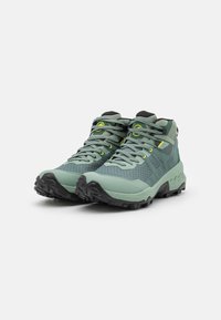 Scarpe da trekking a taglio medio in verde chiaro e verde scuro, caratterizzate da mesh testurizzato, suole robuste e un sistema di allacciatura con accenti gialli.