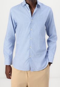 Hombre vestido con una camisa de cuadros pequeño en azul claro, de manga larga y pantalones beige, con una mano en el bolsillo.